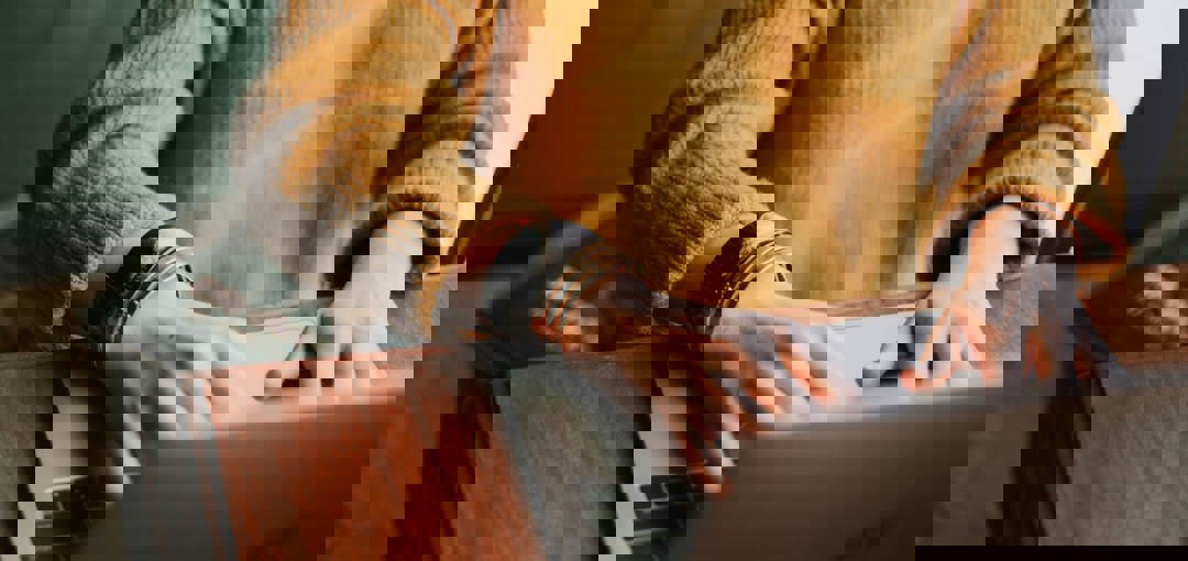 Een vrouw typt geconcentreerd op haar laptop, met een neutrale achtergrond en een focus op haar handen en het toetsenbord.