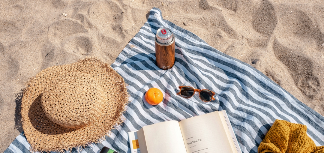 Strandpicknick met gestreepte handdoek, strohoed, geopend boek, zonnebril, camera, mandarijn en koelblikje in de zon op het zand.