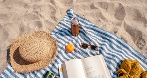 Strandpicknick met gestreepte handdoek, strohoed, geopend boek, zonnebril, camera, mandarijn en koelblikje in de zon op het zand.